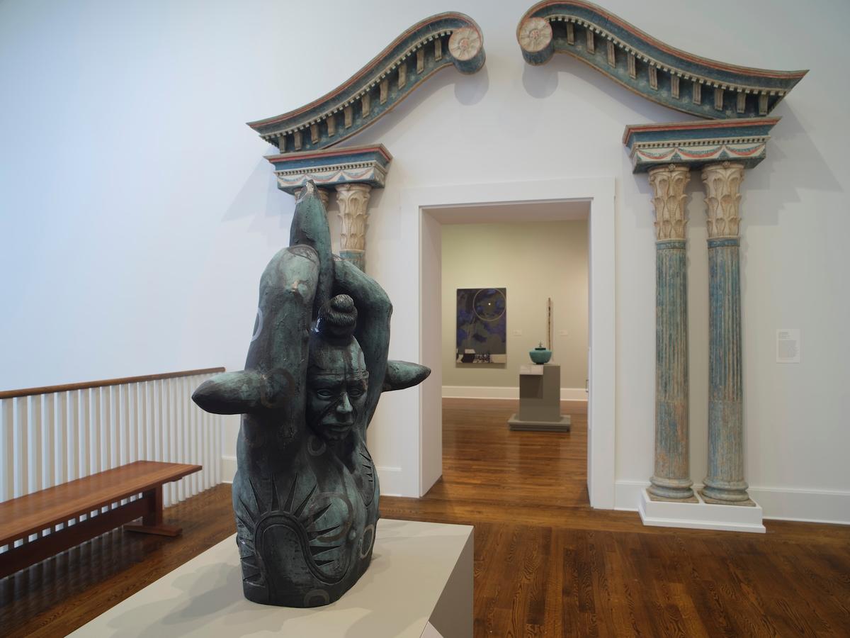 Figurative sculpture on pedestal in front of decorative architectural archway with a view into adjacent gallery.