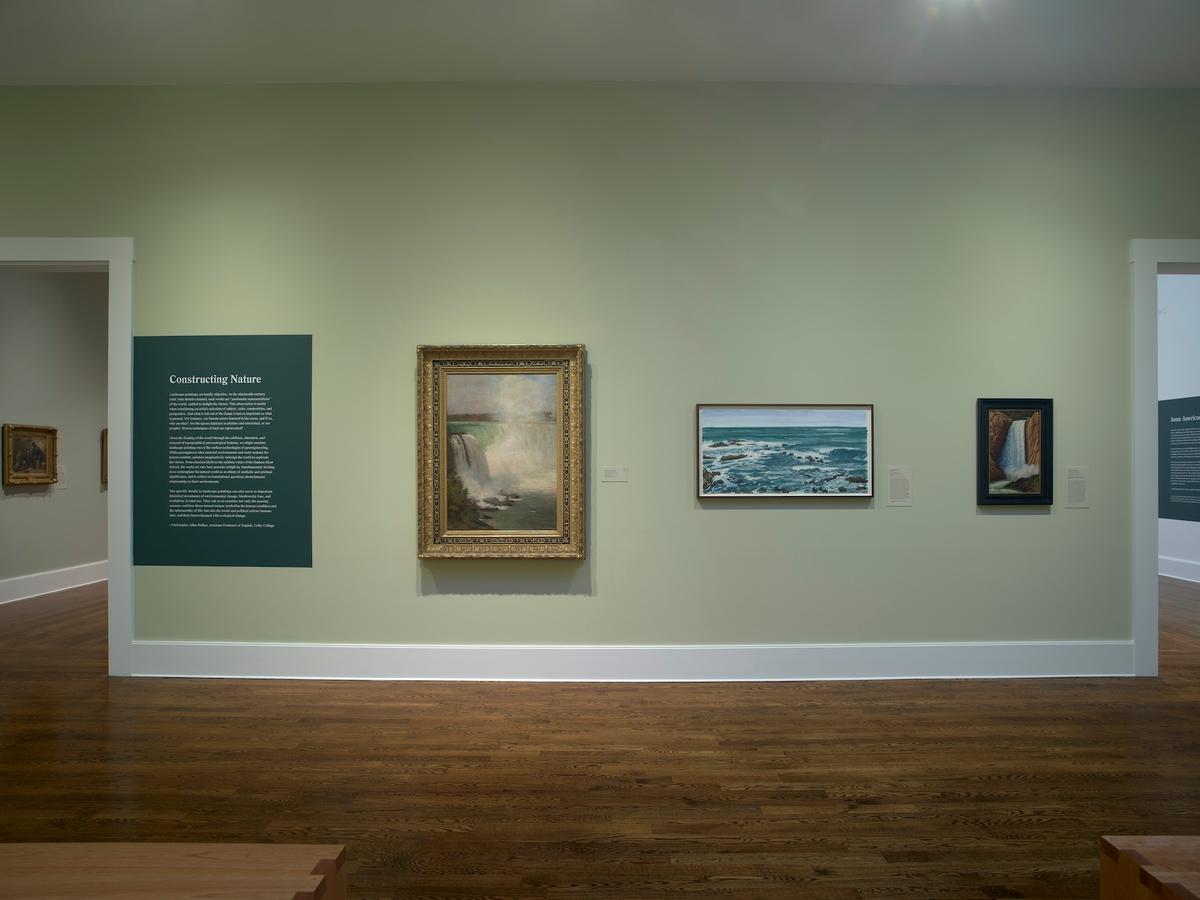 Wall text titled “Constructing Nature” beside three framed landscape paintings with water on a green wall.