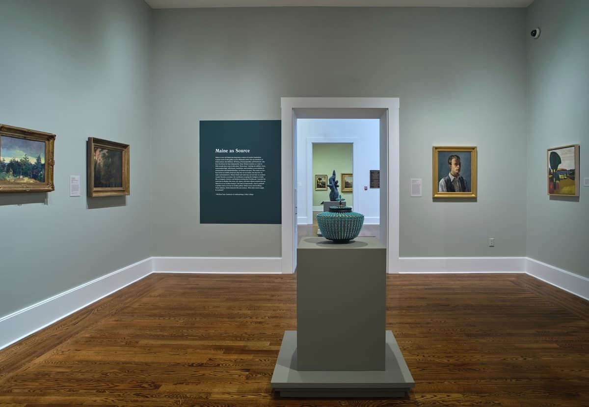 Contemporary basket on pedestal in front of wall text titled “Maine as Source,” with paintings on surrounding walls.