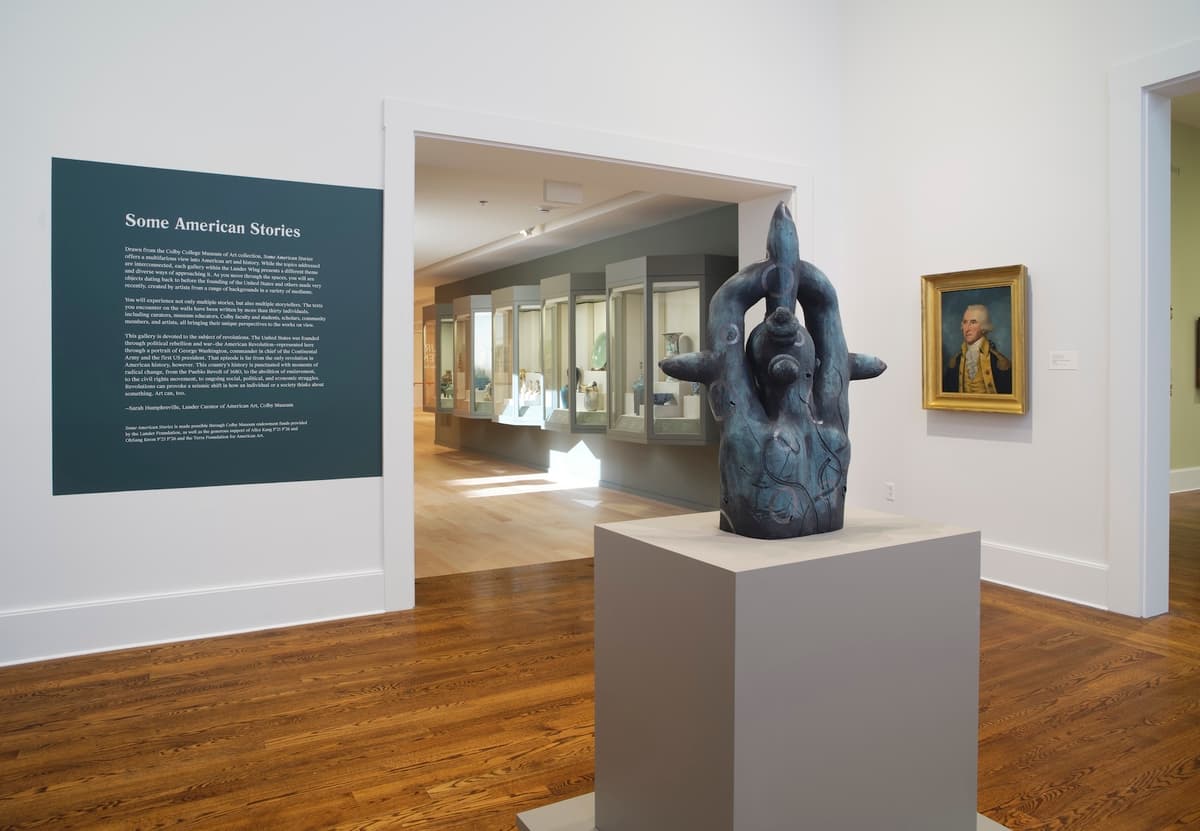 Large ceramic figurative sculpture on pedestal beside wall text and a painting of George Washington, with display cases in the background.