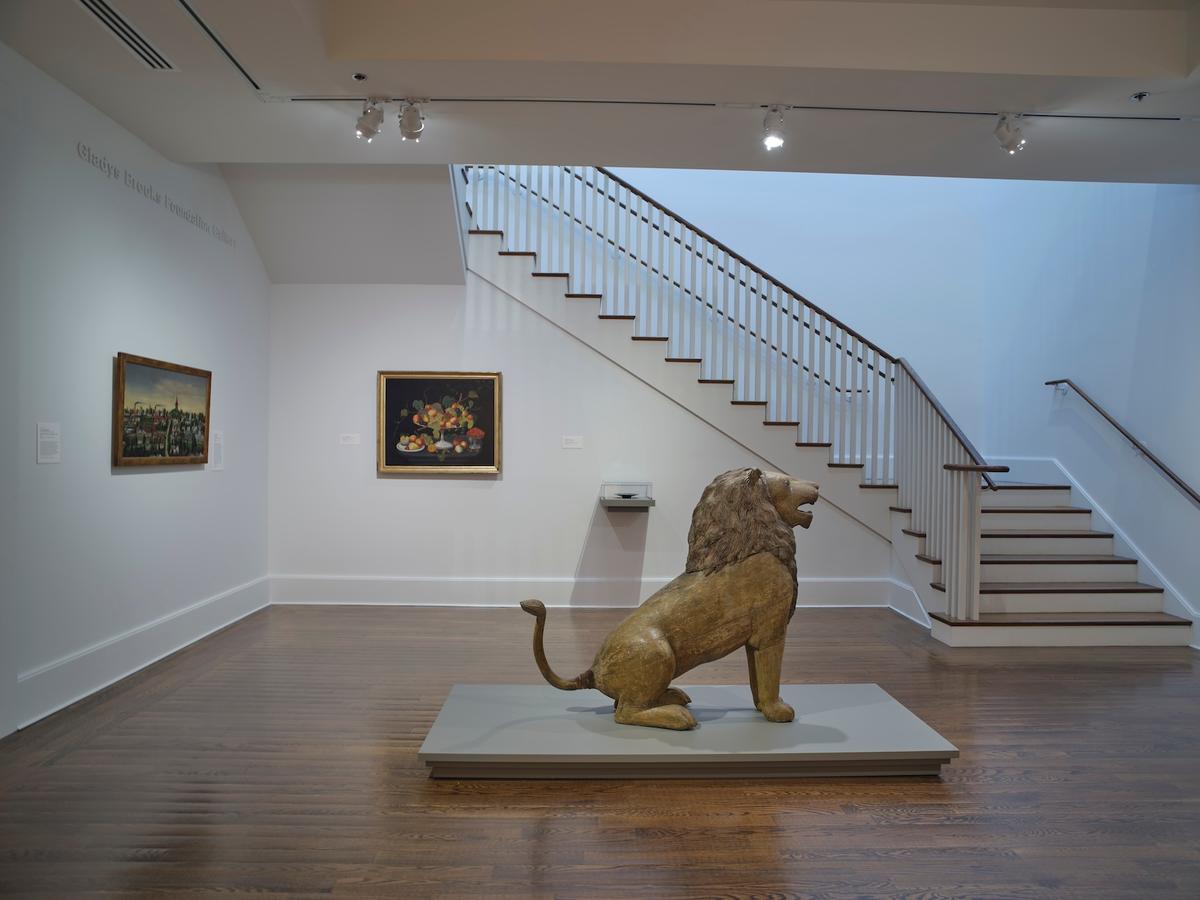 Carved lion sculpture on platform with framed paintings and a staircase in the background.
