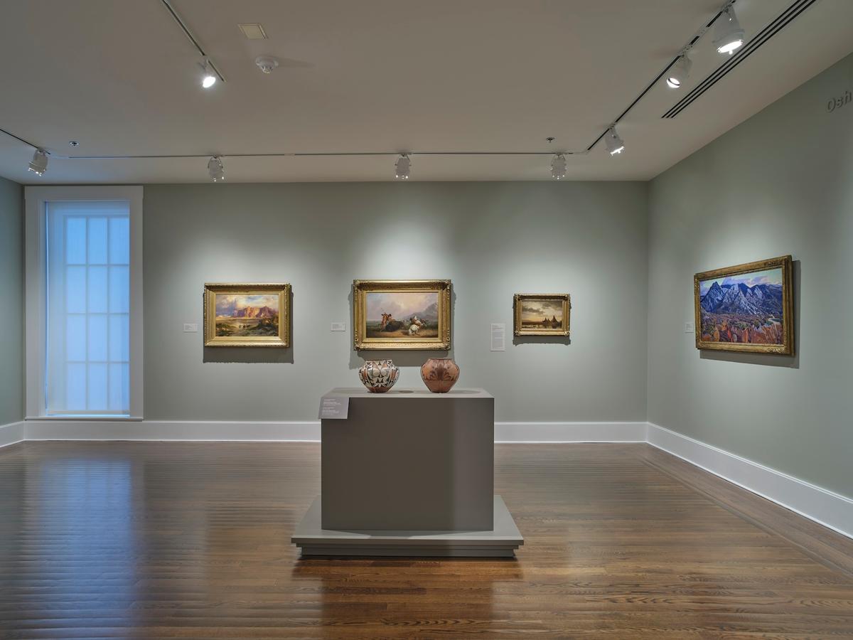 Two ceramic vessels on pedestal surrounded by framed landscape paintings in green-walled gallery.