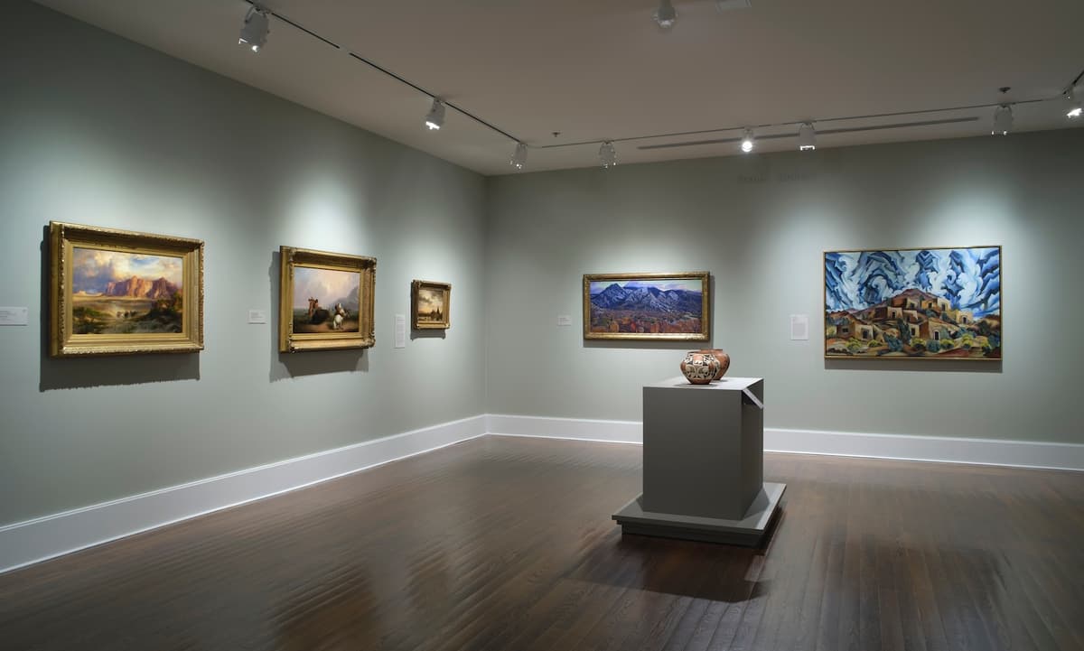 Framed landscape paintings and a ceramic vessel on pedestal in a green-walled gallery.