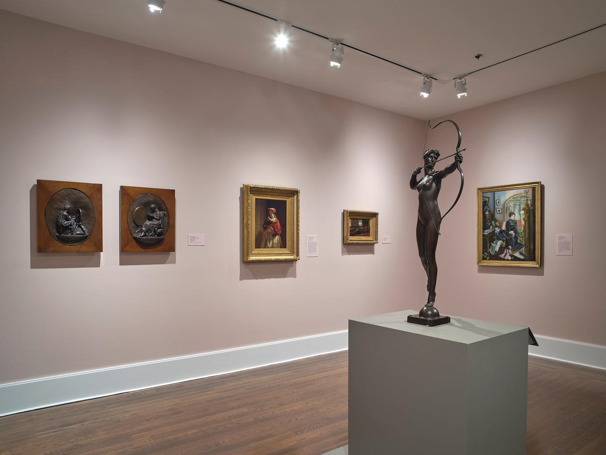 Bronze sculpture of a woman with bow in front of framed paintings and reliefs on pink walls.