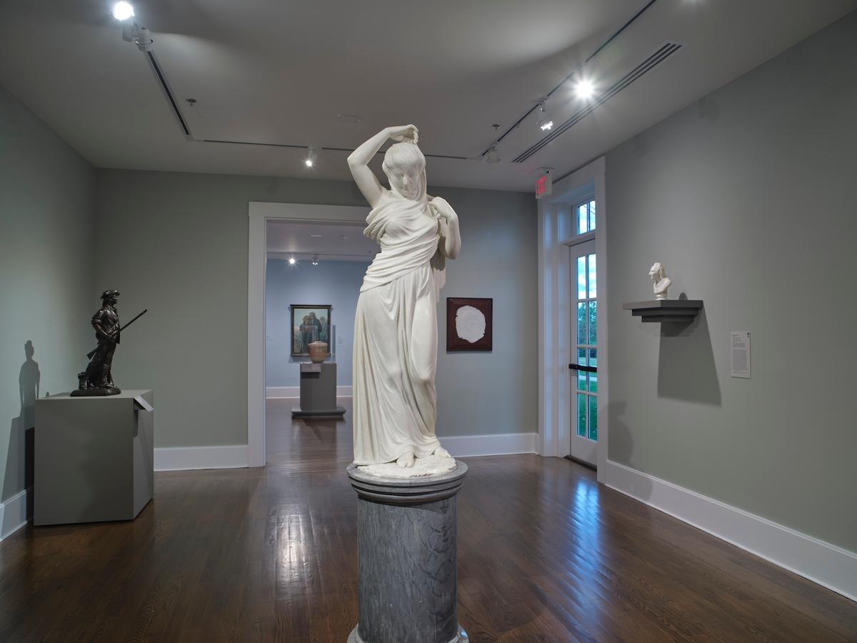 White marble sculpture of a veiled woman on a pedestal in a gallery with additional sculptures and framed works.