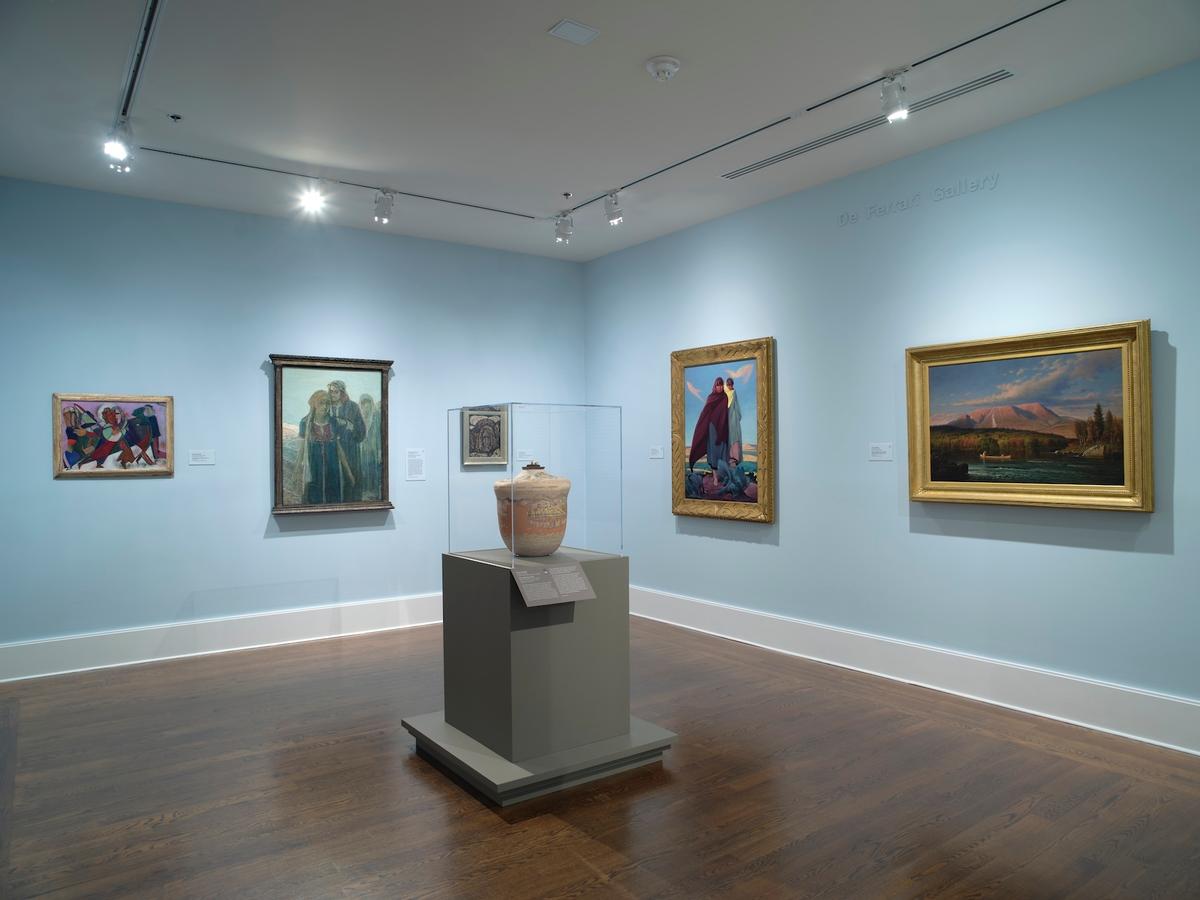 Gallery with blue walls displaying a ceramic vessel on a pedestal and five framed hanging artworks.
