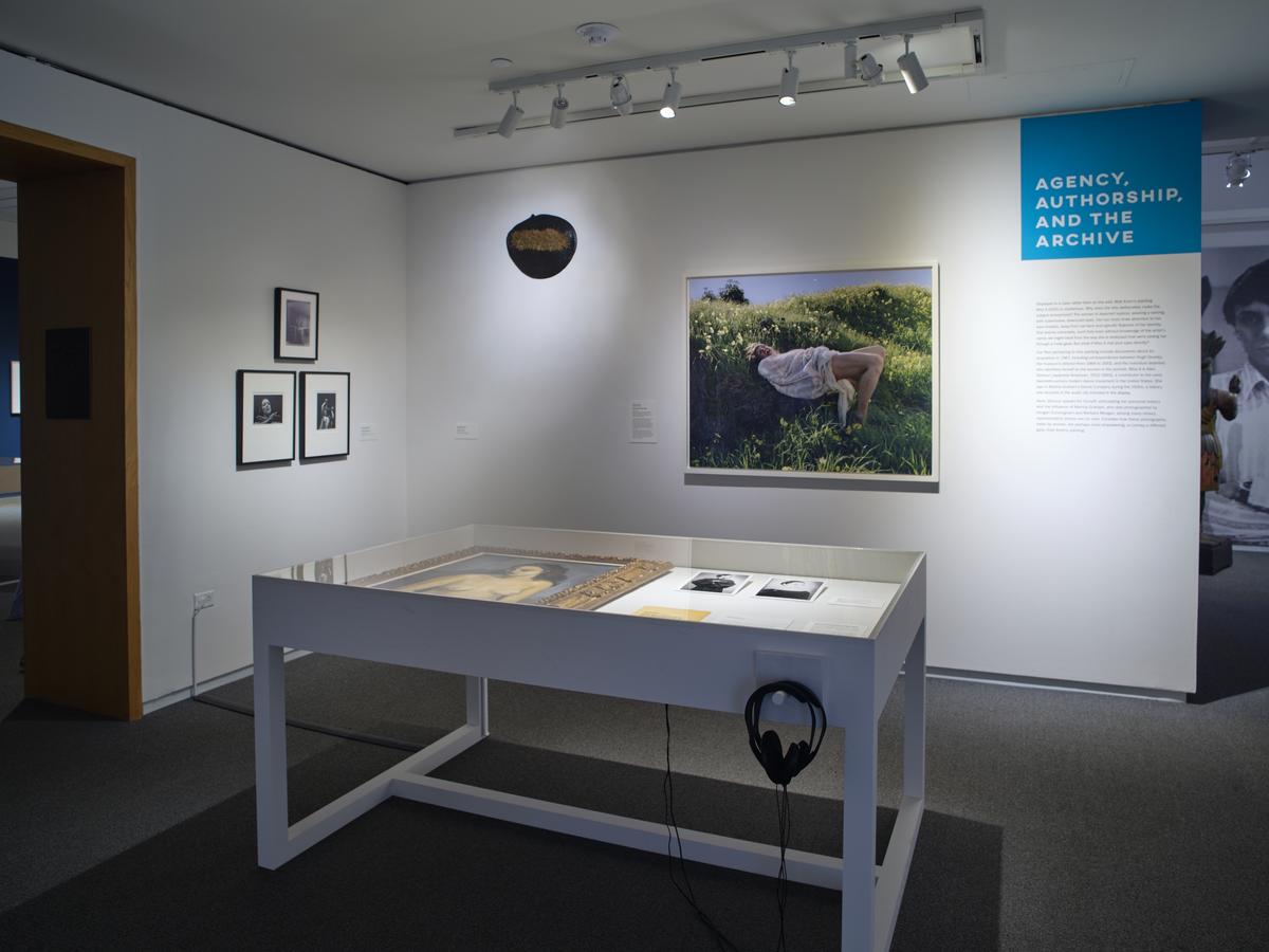 A vitrine with a framed painting and ephemera in the foreground; additional works of art hung on a wall behind including one work depicting a figure lying in tall grass.
