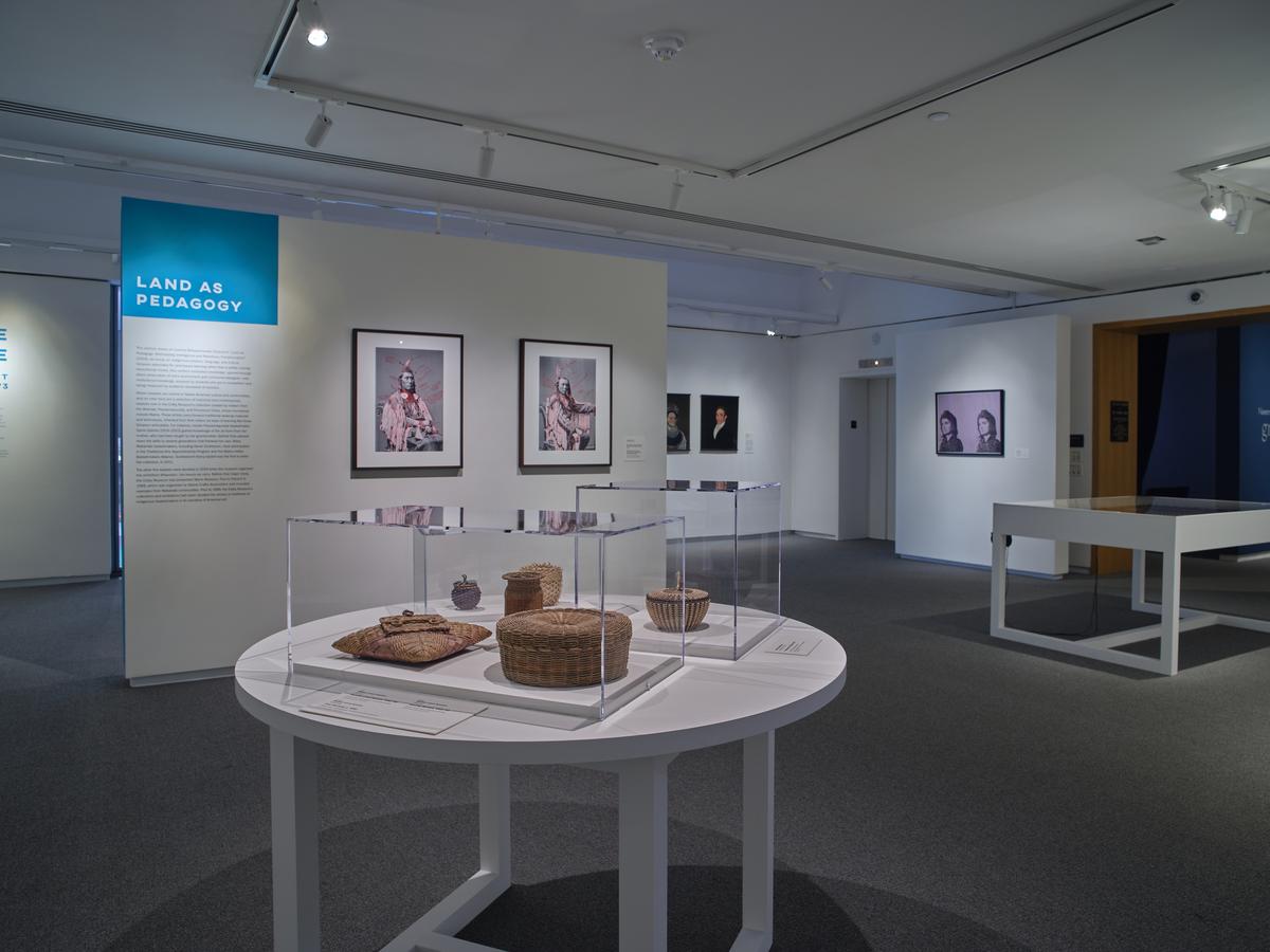 Woven baskets and objects displayed on a round white table, with framed portraits and exhibition text in the background.
