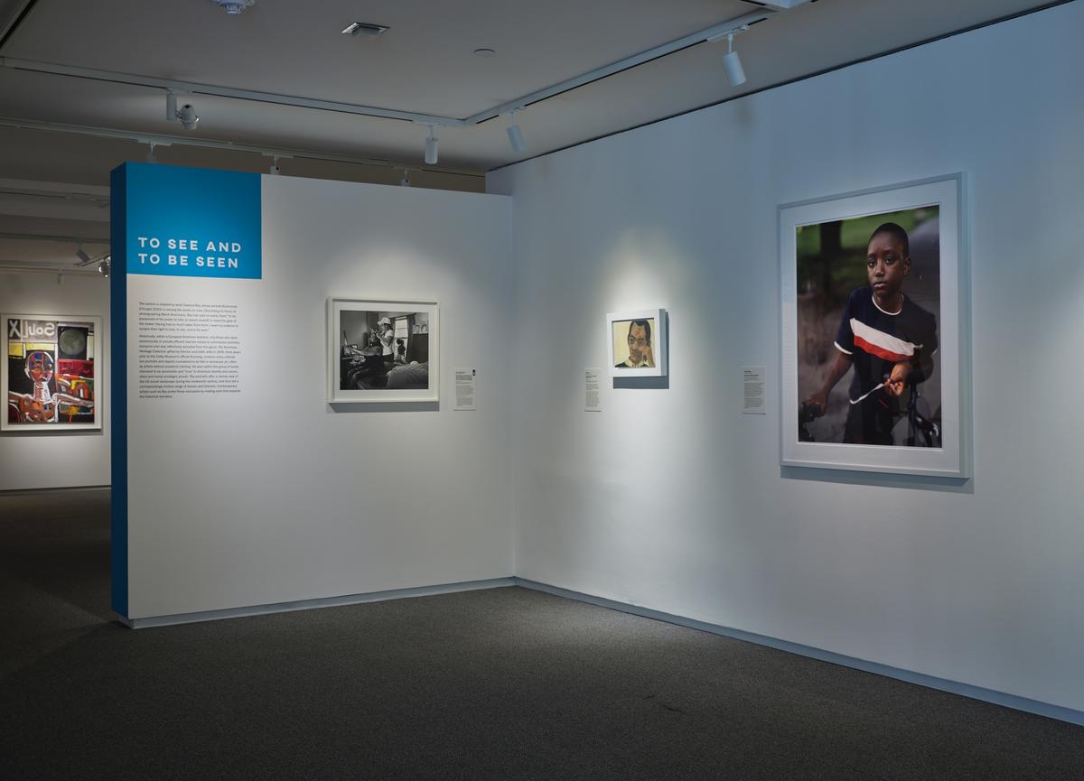 Three framed works of art, each depicting a human subject, next to wall text and under directional lighting in a white-walled gallery corner.