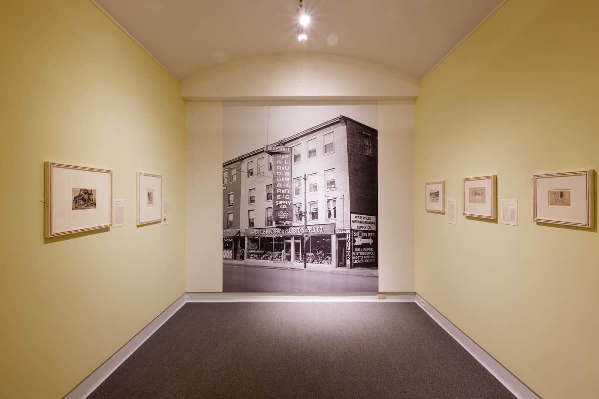 A large black-and-white archival photograph of a Waterville city building centered in a corridor; the adjacent walls each displaying works on paper.