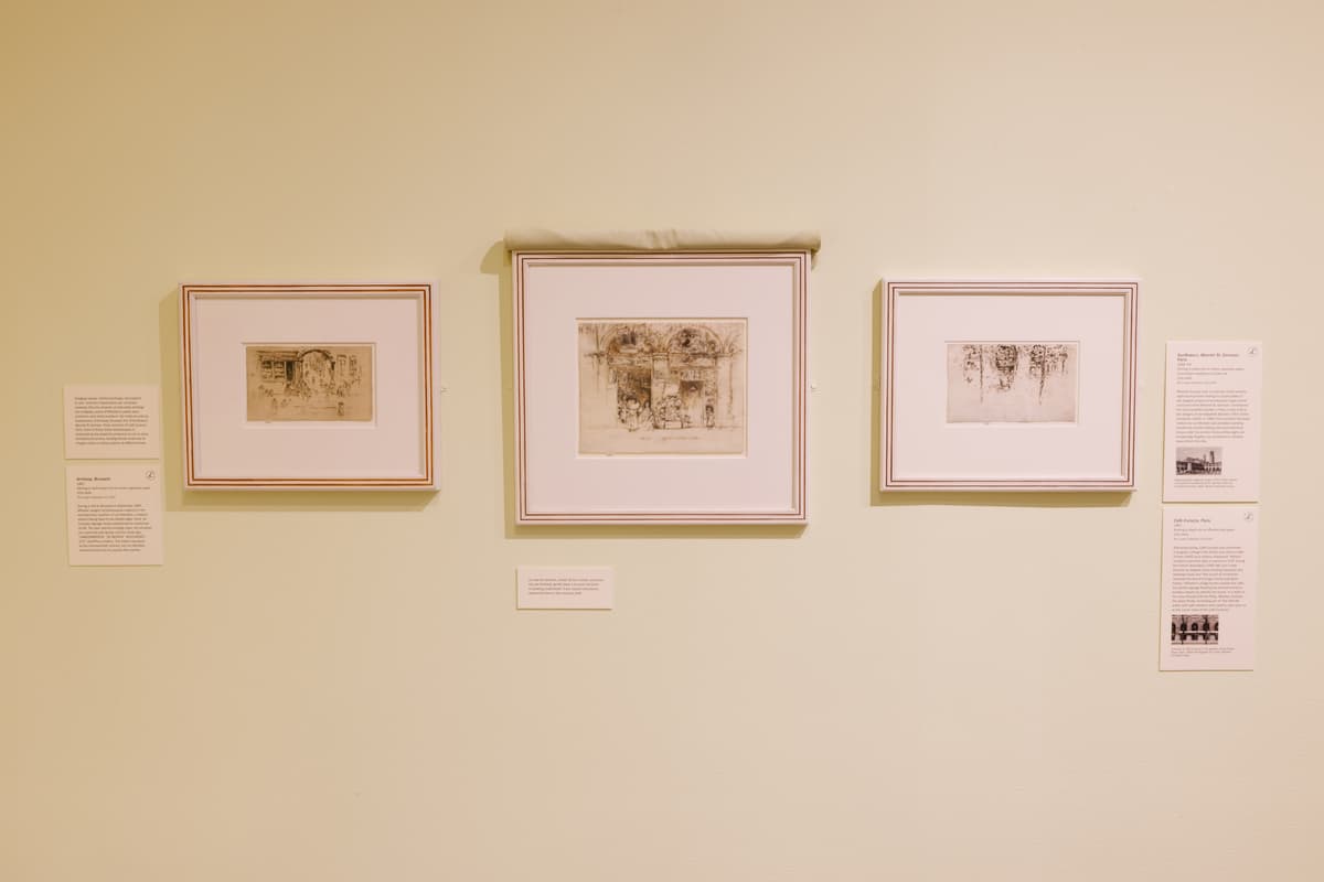Three framed etchings displayed in a row with wall labels and interpretation texts on a pale yellow background; the center work has a piece of fabric rolled up to reveal it, resting on the top of the frame.