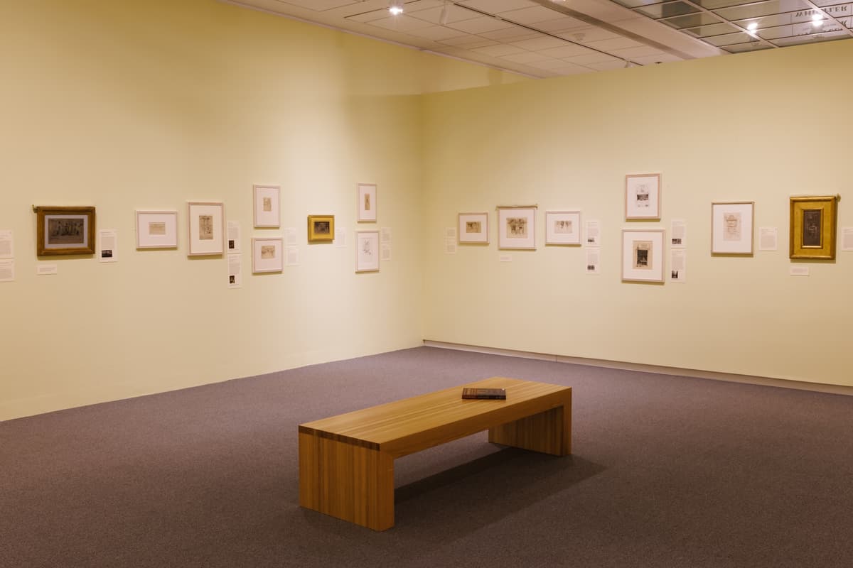 Corner of gallery with an array of framed etchings on two adjacent pale yellow walls; a wooden bench is at the center with a book resting on top.