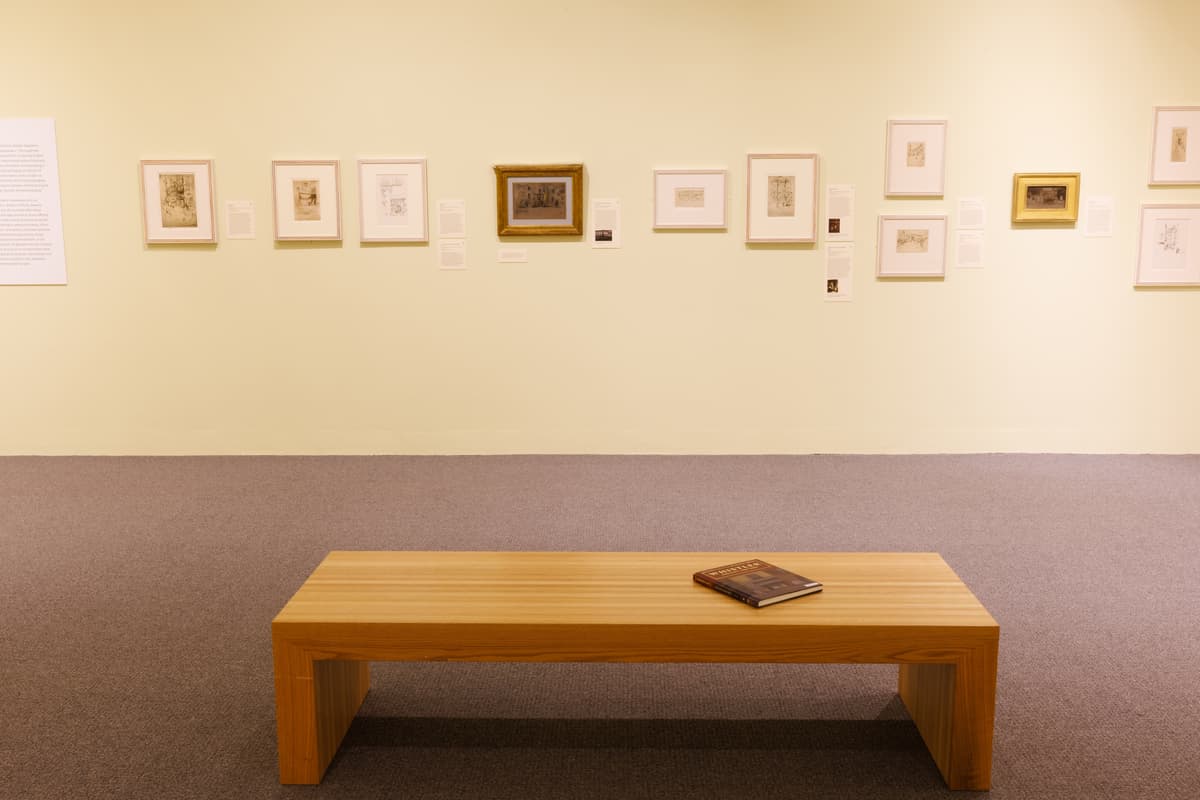 Long wall of framed etchings in varied sizes and tones, with a wooden bench in front.