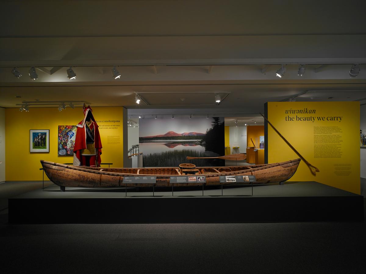 Birchbark canoe displayed in a gallery with mannequin in red regalia, yellow interpretive panel, and landscape mural