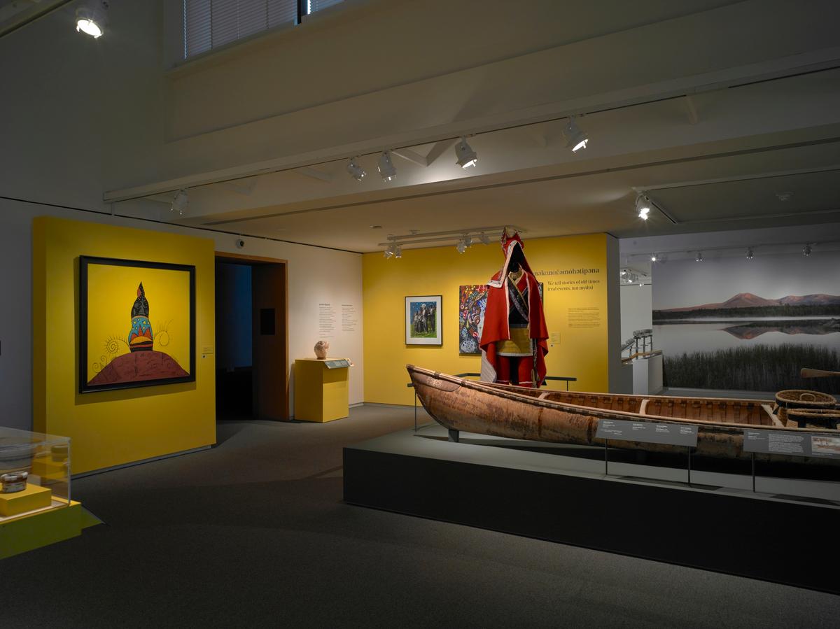 Birchbark canoe and mannequin in red regalia on display in a gallery with yellow walls and framed artwork
