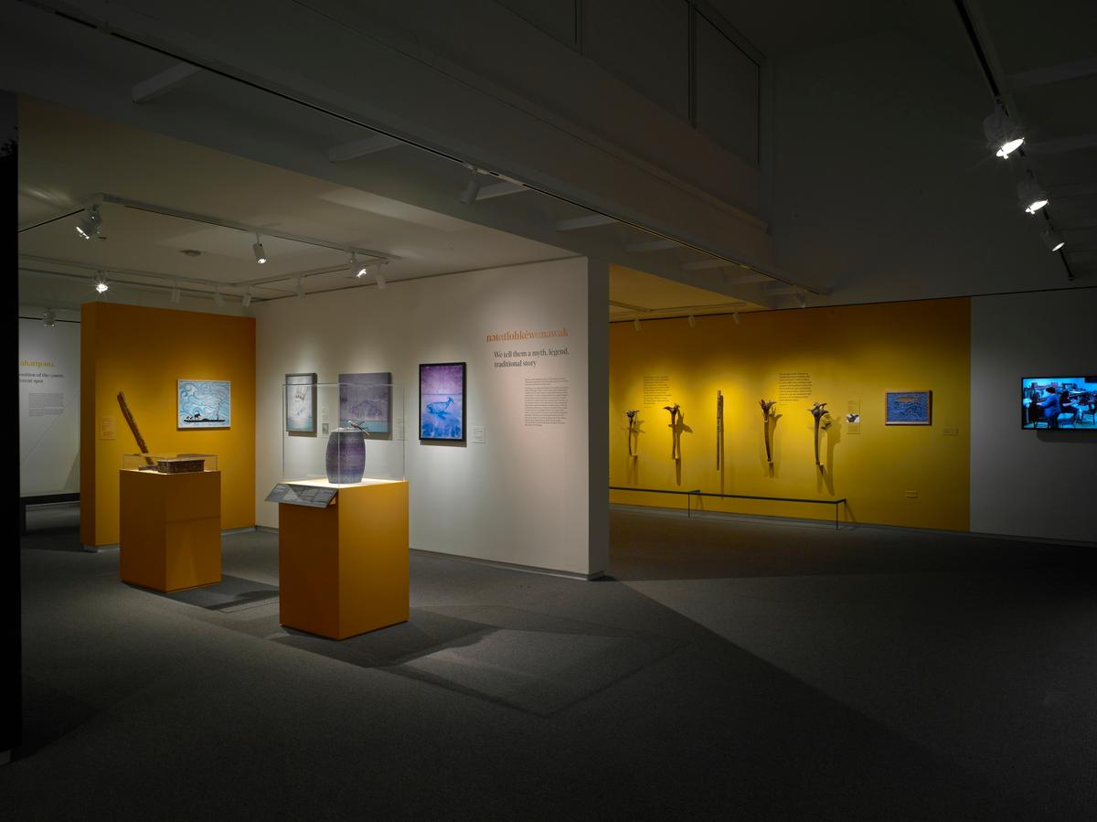 Gallery view with ceramic vessels on yellow pedestals, framed artworks on white walls, and carved paddles on a yellow wall in the background