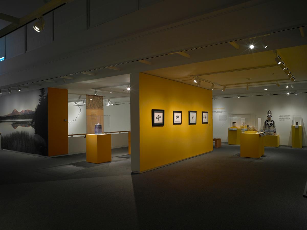 Gallery view with framed artworks on a yellow wall, ceramic vessels on pedestals, and a mannequin in a beaded garment