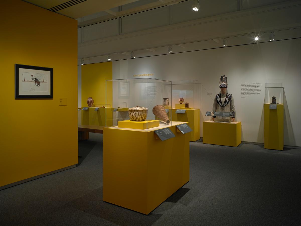 Baskets, pottery, and a beaded garment displayed in cases on yellow pedestals in a gallery with framed art on the wall
