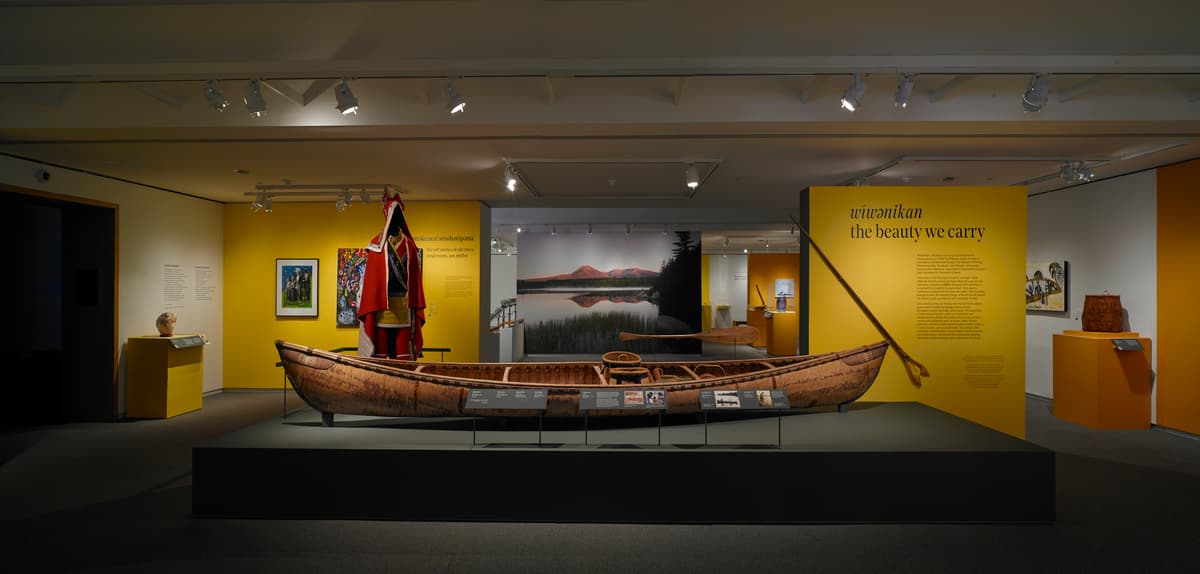Birchbark canoe displayed in a gallery with mannequin in red regalia, yellow interpretive panel, and landscape mural