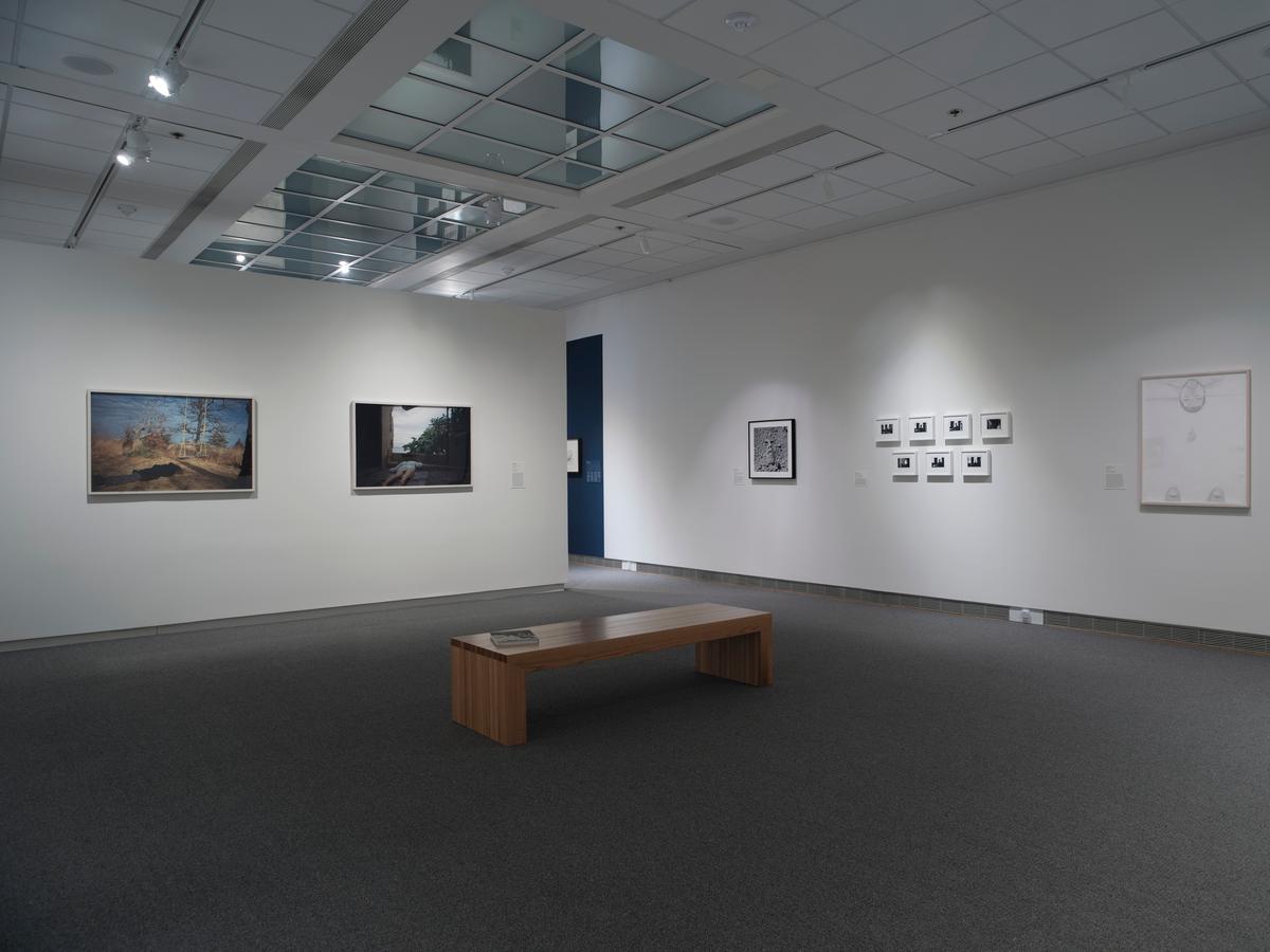 Several framed artworks spanning medium on white gallery walls with a wooden bench in the center of the room.