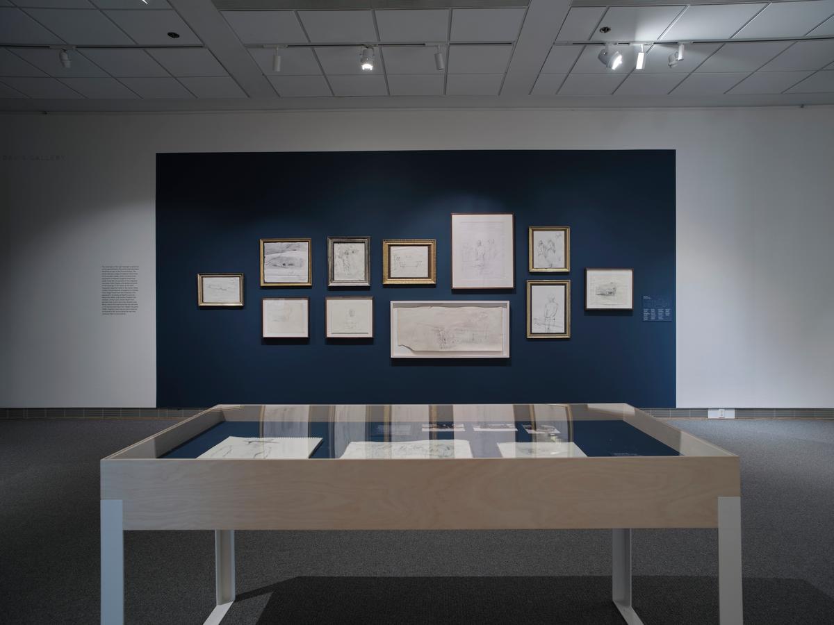 Cluster of framed drawings on navy wall above wooden display case in gallery setting.