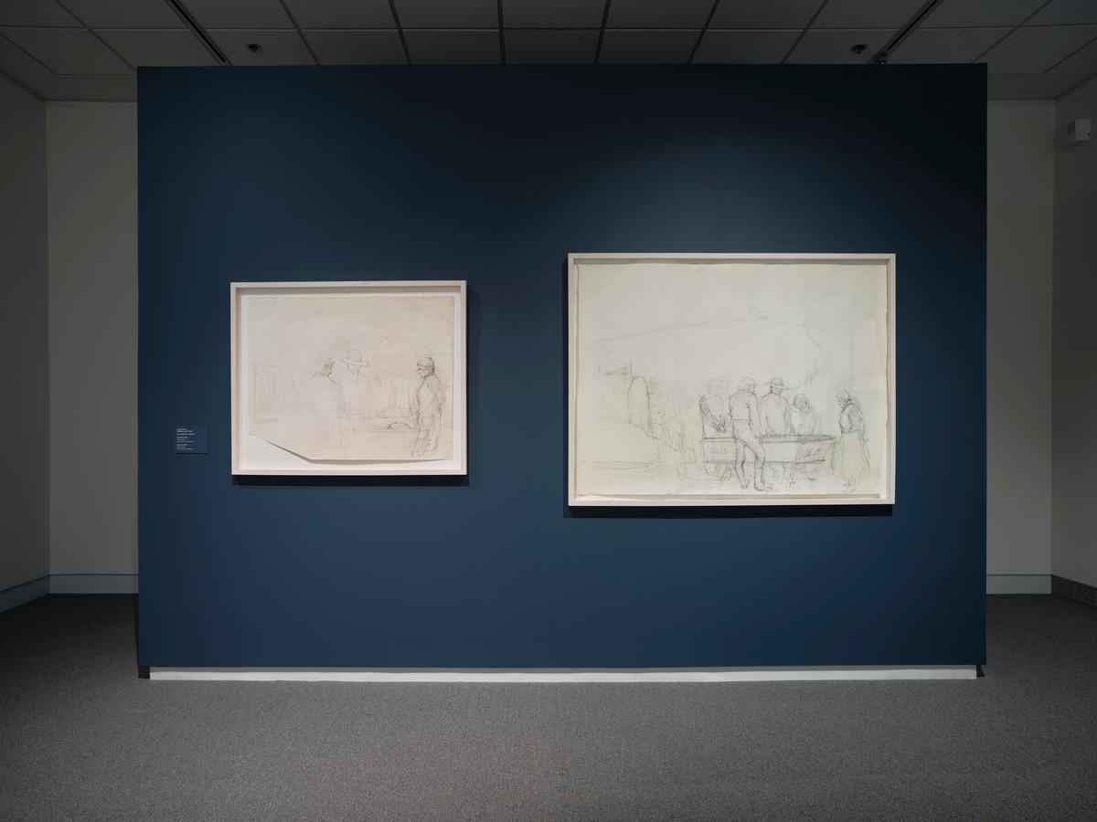 Two framed drawings, each showing a group of figures, hung on a dark blue gallery wall with gray carpeted floor.