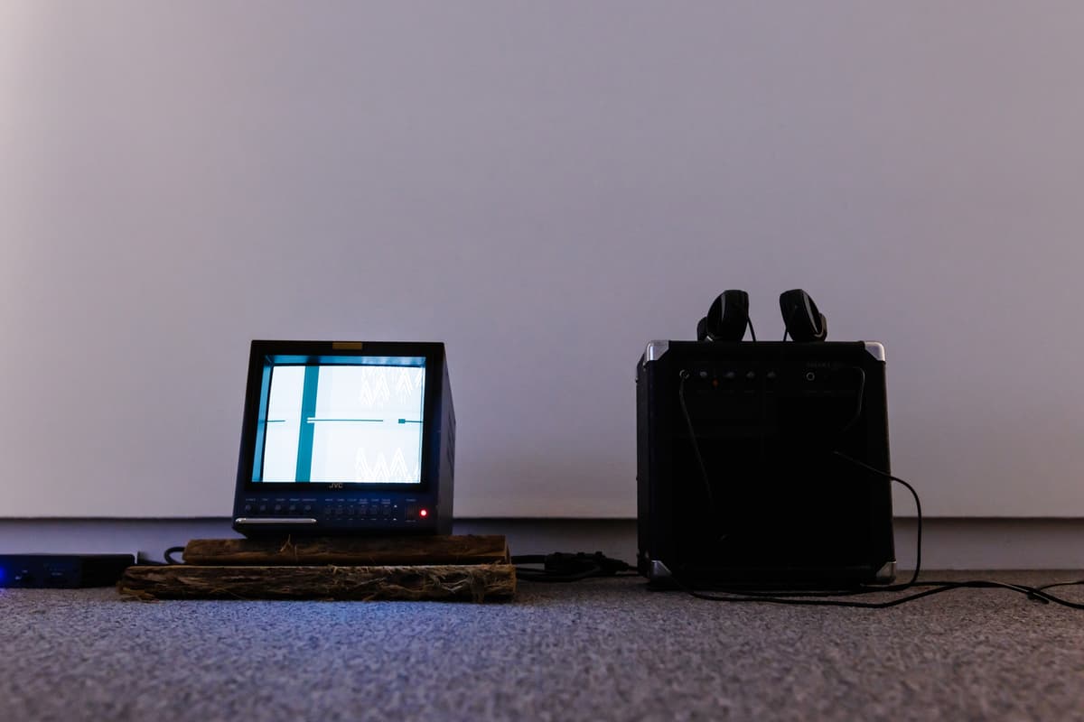 A monitor, next to a black speaker with headphones resting on top, against a white gallery wall.