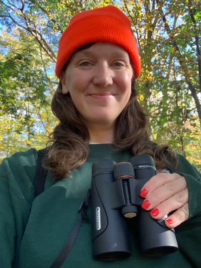 A selfie of a woman wearing a bright orange beanie and holding a pair of binoculars.