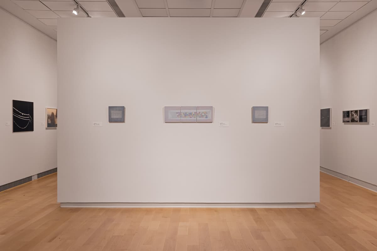 Installation view of a wide gallery wall featuring several diverse, spaced-out artworks, including small paintings and photographs. From left to right, a dark square photograph, a blurred color photo, two small abstract square works, a central rectangular piece with small colorful elements, and two small works on the far right.