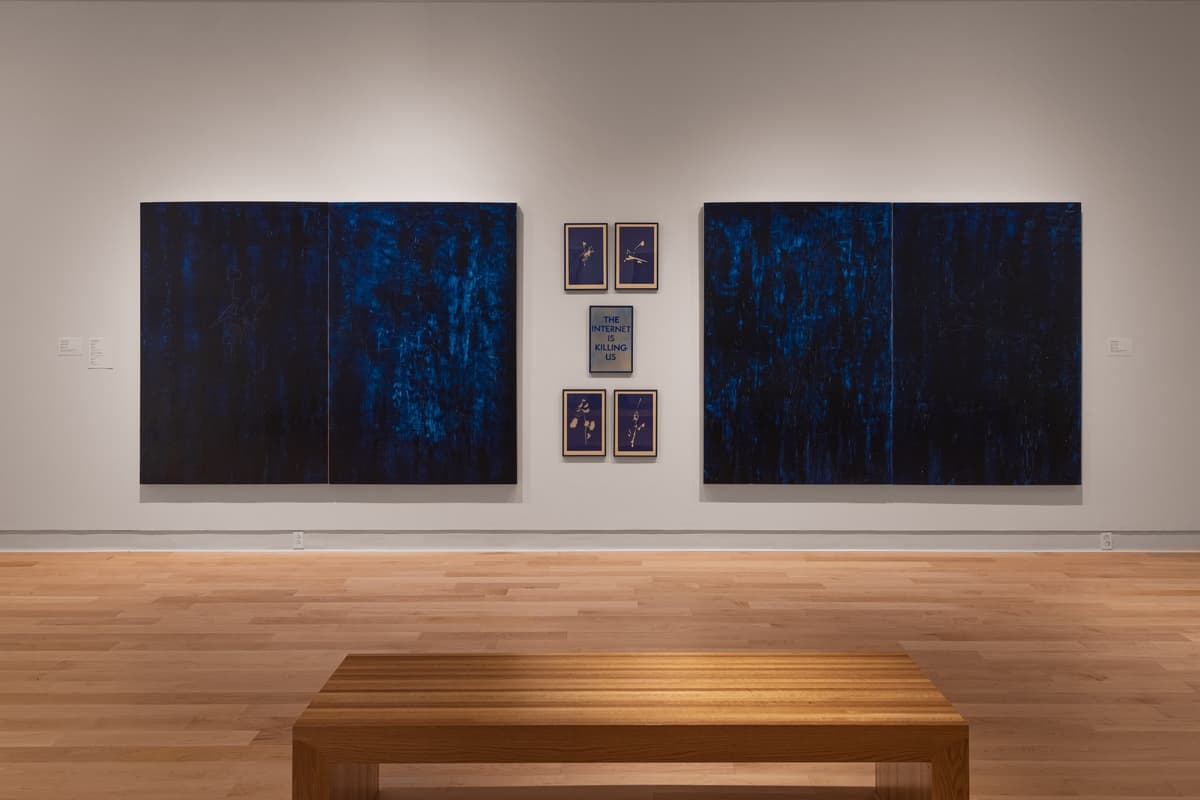 Installation view of the "2025 Faculty Biennial," featuring two large, dark blue, abstract indigo diptychs flanking a central grid of six smaller framed works, one of which contains the text "THE INTERNET IS KILLING US." A light wood bench is in the foreground.