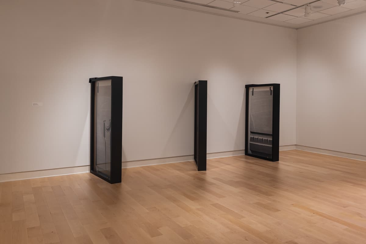 Installation view of three sculptural works resembling dark, framed window or door frames standing in the corner of the gallery. The central piece is turned sideways, presenting a thin black profile. The work suggests an exploration of structure or barriers.