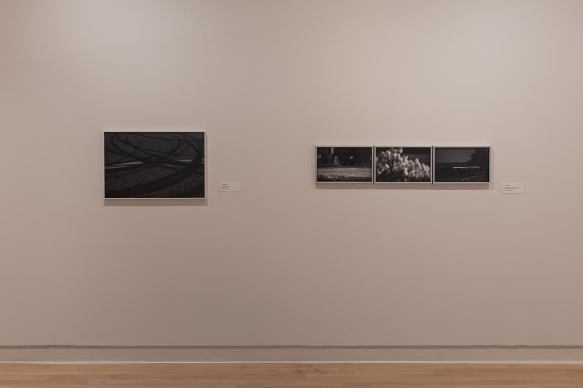 Installation view of two sets of black-and-white photographs: on the left, a single horizontal image showing tire tracks or skid marks on a dark surface; on the right, a triptych of three small horizontal images depicting indistinct outdoor scenes, possibly including foliage or figures.