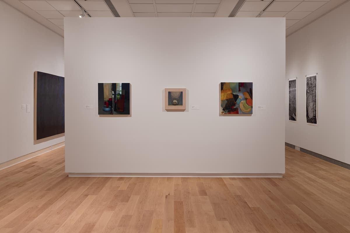 Installation view of a central gallery wall displaying three square paintings: a dark interior still life on the left, a small abstract piece with a light frame in the center, and a colorful abstract still life on the right. A large, dark artwork is visible on the wall to the far left.