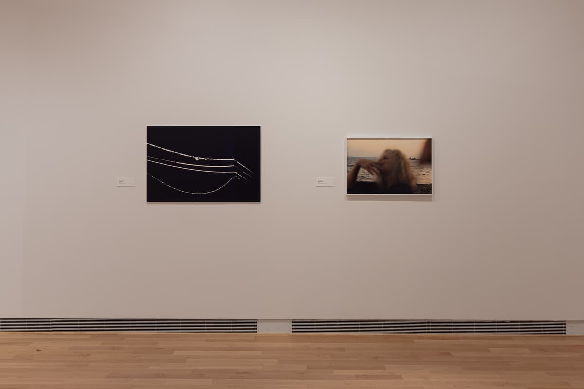 Installation view of two framed artworks: on the left, a square, dark photograph featuring thin, arcing white lines resembling strings of lights or reflections; on the right, a square, color photograph of a blonde figure, blurred and in profile, against a sunset-colored background near water.