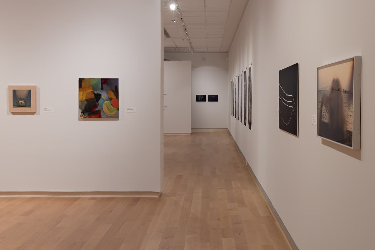 Installation view showing a corner of the gallery with a hallway extending into the distance. On the near walls are several artworks: a small still life painting and a colorful, abstract still life painting on the left wall, and two square photographs (dark abstract and blurred figure) on the right wall.