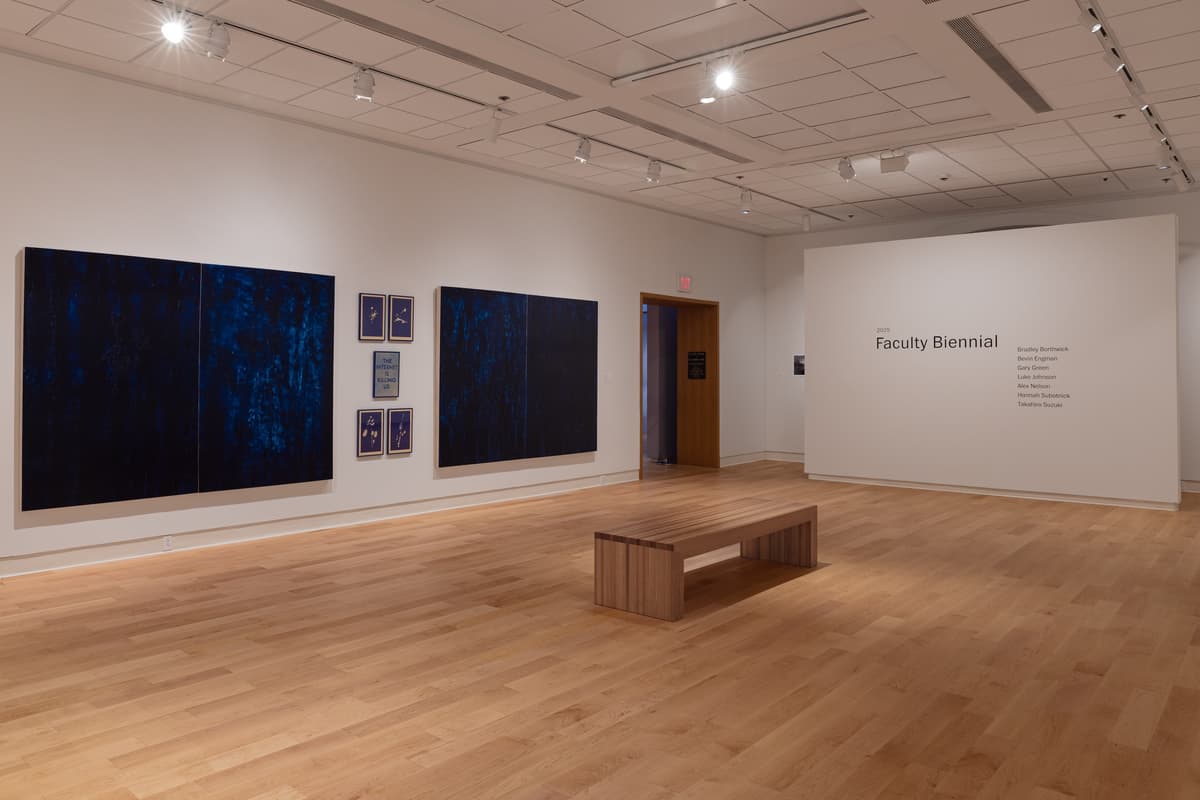 Installation view of the "2025 Faculty Biennial" exhibition. The center wall displays two large, dark blue abstract cyanotype diptychs flanking a central grid of six smaller framed works, one of which contains the text "THE INTERNET IS KILLING US." To the right is a white wall with the exhibition title and a list of faculty artists' names displayed. A light wood bench sits in the middle of the floor.