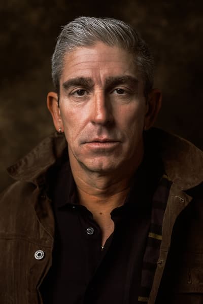 Portrait of a man directly facing the camera with a dark background. The man wears a black shirt and brown jacket.