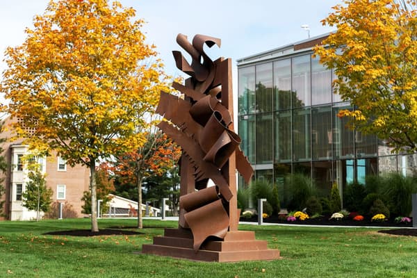 A monumental, abstract outdoor sculpture by Albert Paley made of rusted corten steel. The large, reddish-brown artwork consists of towering, interlocking, and curling ribbons of metal, set on a tiered base on a grassy lawn. The sculpture is framed by trees with golden autumn foliage, with modern glass and brick academic buildings in the background.
