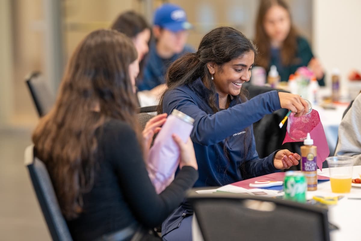 A medium-angle shot shows two young women seated at a table, participating in an art activity with a group of people partially visible in the blurred background. The woman on the left, with long dark hair, wears a black top and holds a pale purple thermos or insulated bottle. The woman on the right, smiling and looking down at her work, has long dark hair pulled back and wears a dark blue quarter-zip top with "COLBY CENTRAL" visible on the chest. She's holding a small silver can and a piece of magenta paper or fabric, appearing to be cutting or decorating. On the table are various craft supplies including a bottle of Tacky Glue, a pair of scissors, a green and white can of soda, and a cup with an amber drink. The background is a brightly lit, modern-looking indoor space.