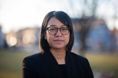 A woman with dark, shoulder-length hair looks directly at the camera. She wears brown-framed glasses, and her expression is neutral. She is dressed in a dark blazer over a dark top. The background is softly blurred with warm, natural light, showing hints of bare trees and residential structures, suggesting an outdoor setting during a sunny day.
