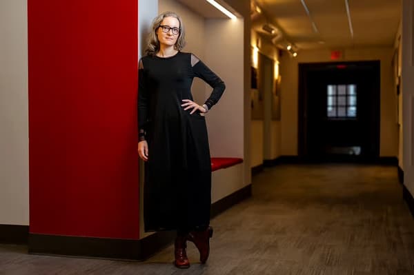 A woman with gray hair and glasses, wearing a long-sleeved black dress and reddish-brown boots, stands with one hand on her hip in a hallway with a red accent wall.