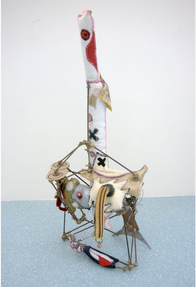 A vertical photograph of a mixed-media sculpture resembling a primitive or deconstructed figure, standing on a pale blue floor against a plain white wall. The structure is supported by thin brass or copper rods and wires, and it incorporates various stitched fabric elements, including a tall, cylindrical white form with red patches at the top, and smaller, stuffed, irregularly shaped pieces with exposed zippers, buttons, and cross-stitching, giving it a Frankenstein or rag-doll aesthetic. Small, circular metal discs resembling cymbals are attached to the upper left side.
