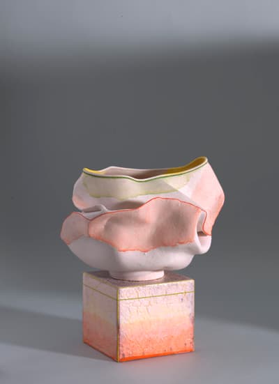 A contemporary, light pink ceramic bowl or vessel, possibly by Warren Buterly, with an irregular, undulating, and layered shape resembling a crumpled flower or piece of fabric. The upper edges are detailed with thin bands of yellow and green stitching or paint. The vessel sits on a small, square pedestal with an ombre orange, pink, and yellow crackled surface, outlined with thin gold trim. The entire piece is set against a solid gray background.