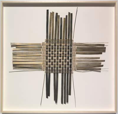 A framed, mixed-media artwork by Julia Frey, consisting of thin strips of natural material woven into a square grid pattern centered on a white background. The weaving extends outward in four directions (up, down, left, right) forming a cross shape. The strips are varied in color, ranging from light beige to dark gray and black, creating a subtle checkerboard effect in the center. Loose, fine strands of thread or fiber extend from the corners of the woven structure.
