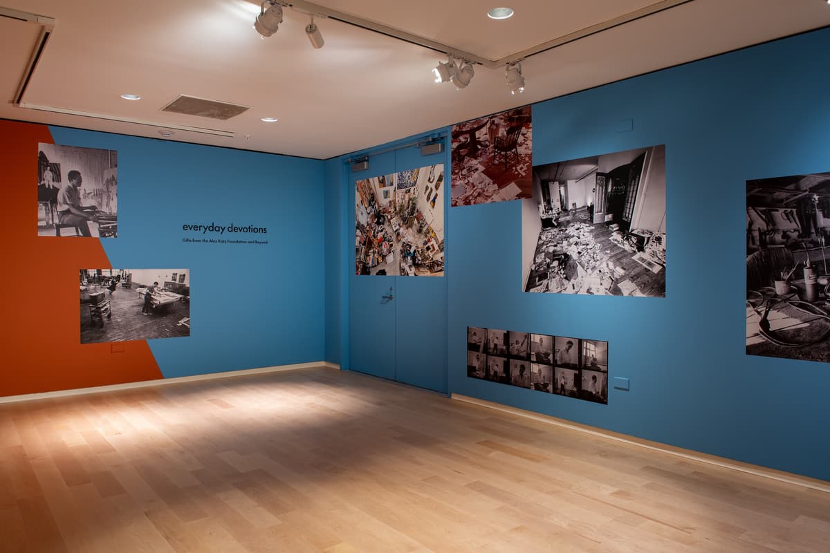 Installation view of a gallery with light wood floors and blue walls. Large-scale black-and-white and color photographs of artist studios are mounted directly to the blue wall.