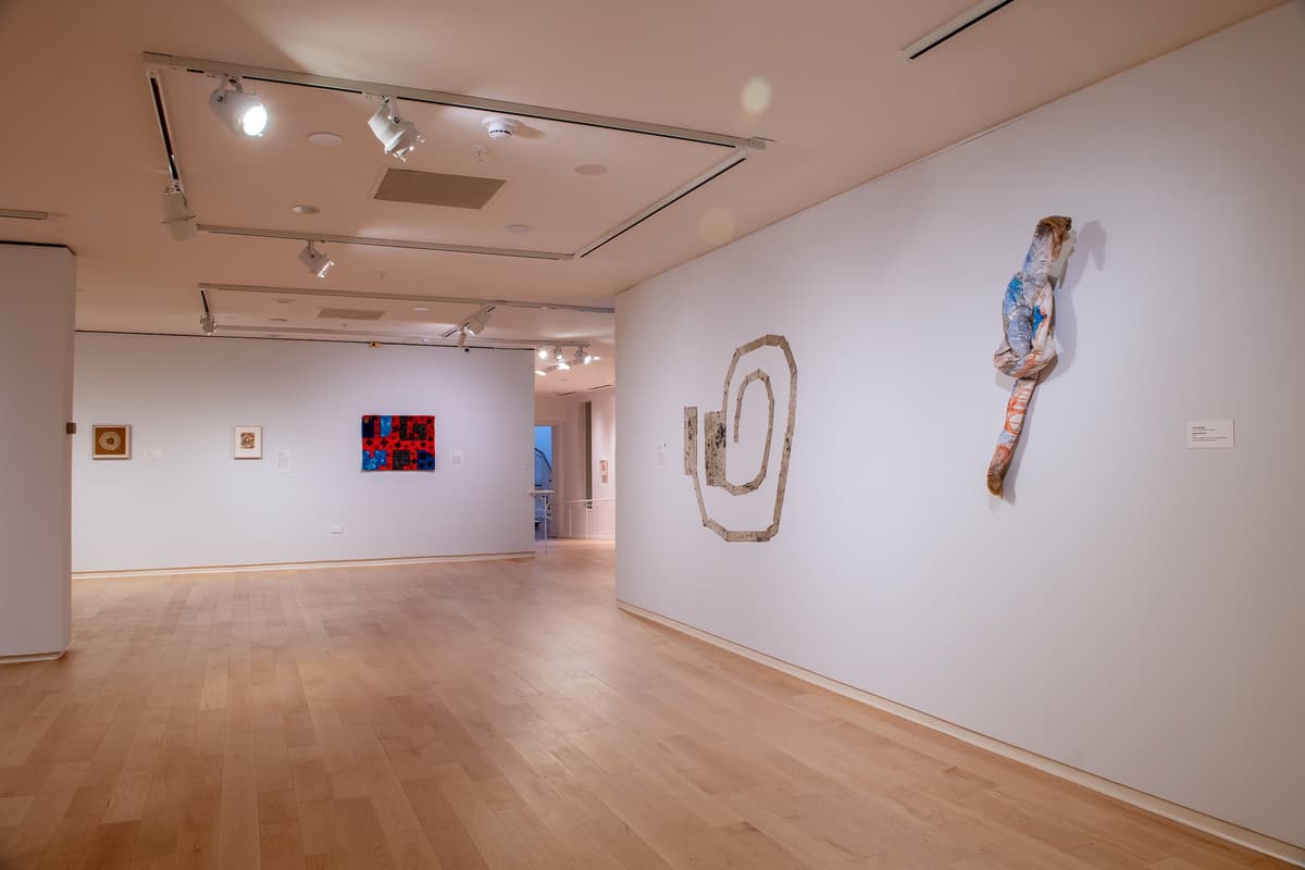An open gallery space with white walls and track lighting. On the right wall, a large, tan, coiled fabric sculpture and a vertical, knotted textile work are displayed.