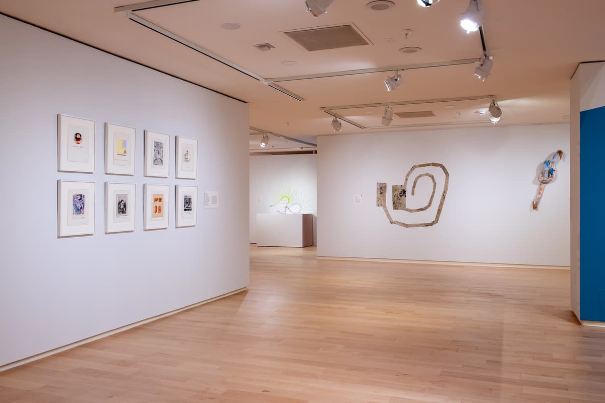 Gallery view featuring a white wall on the left with eight small framed works on paper arranged in two rows. In the background, textile sculptures hang on a far wall.