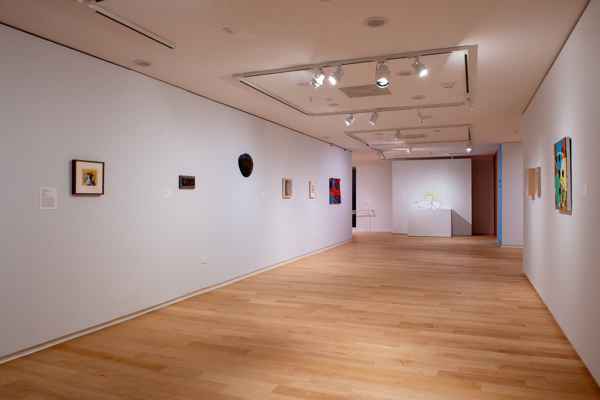 A wide-angle view of a long gallery wall featuring several small, framed mixed-media works and a three-dimensional dark mask-like object. A colorful quilt hangs further down the wall.