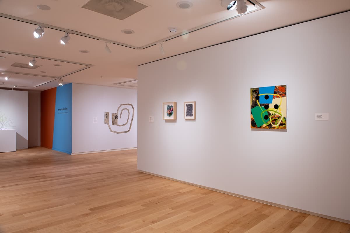 Close-up of a white gallery wall featuring three small framed works on paper. To the right, a square abstract painting with overlapping blue, green, and orange shapes, black circles, and yellow lines.