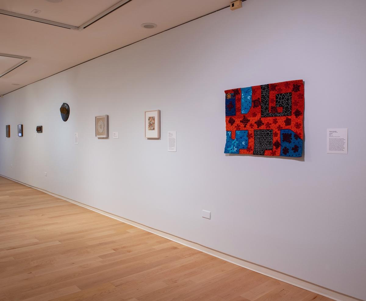 Angled view of a white wall showcasing a colorful, rectangular fabric work with red, blue, and black patterned sections, alongside other small framed artworks.